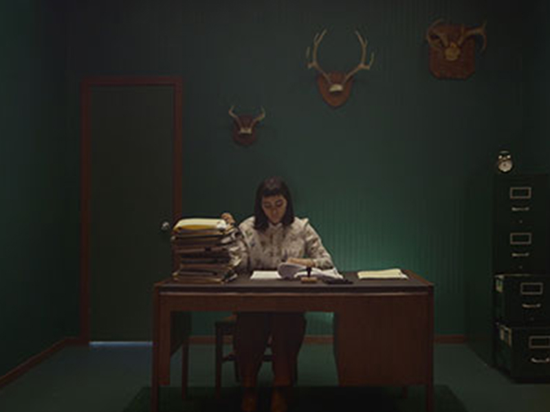 A still from Carol Nguyen’s Facade, a person sits at a dimly lit desk with piles of paperwork