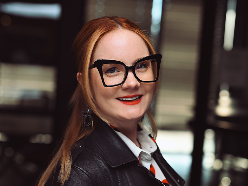 Melissa Tuplin with dark-rimmed glasses standing in profile looking forward wearing a lather jacket with white shirt