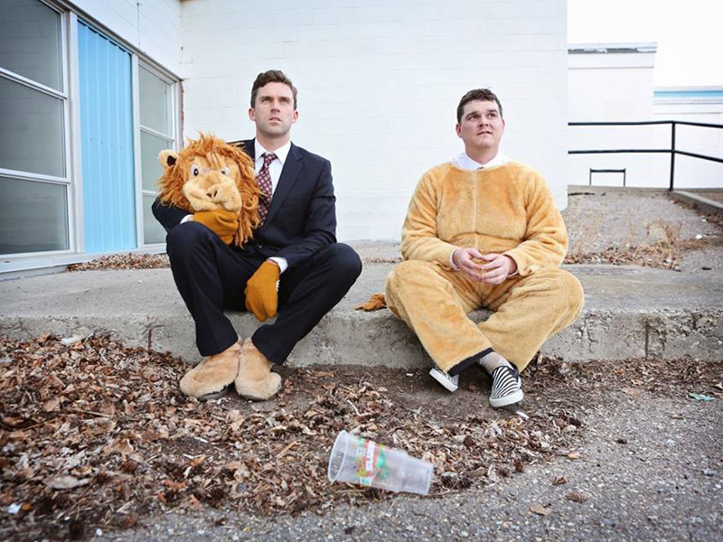 A man in a suit holds a lion mask next to a man wearing its costume