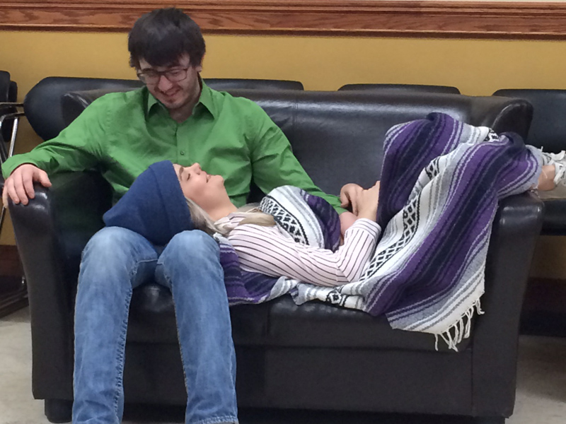 A man sits on a couch as a woman lays across his lap wrapped in a blanket