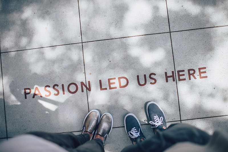 A photo of two people's shoes next to the words Passion Led Us Here on a sidewalk