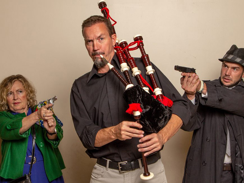 A man and a woman point their guns at a man playing bagpipes