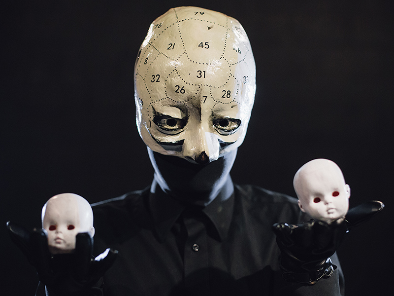 A creepy figure in a mask holds two pale, white and bald dolls