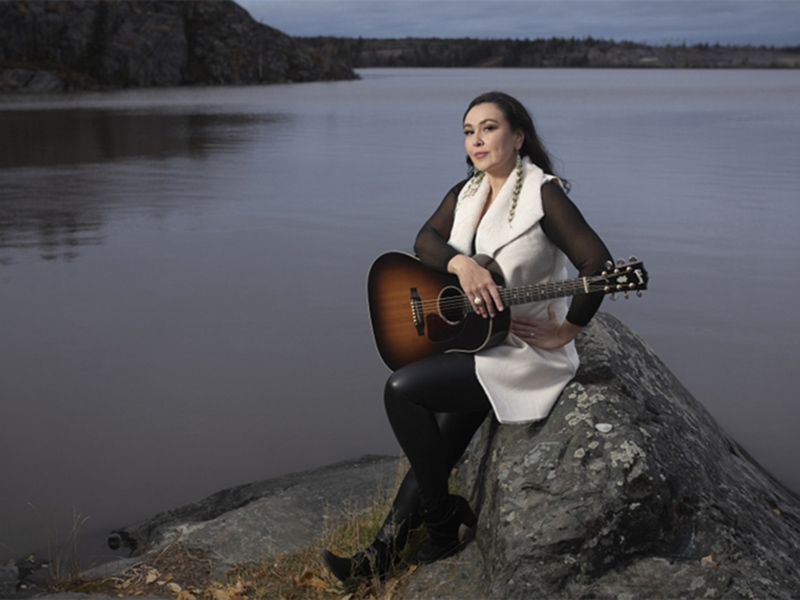 Leela Gilday holds a guitar while sitting next to a pristine lake
