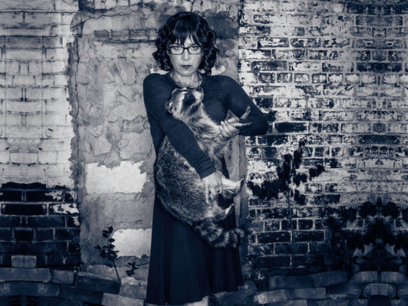 Karen Hines holds a taxidermy racoon against a brick wall