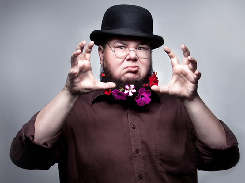 A promotional photo of Shane Koyczan wearing a bowler hat and flowers in his beard