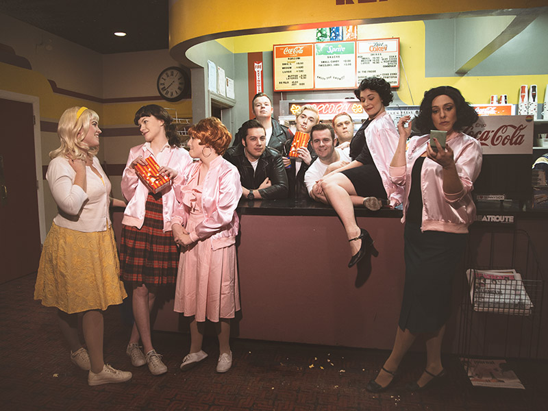 The cast of Front Row Centre's production of Grease stand around the concession at The Plaza Theatre
