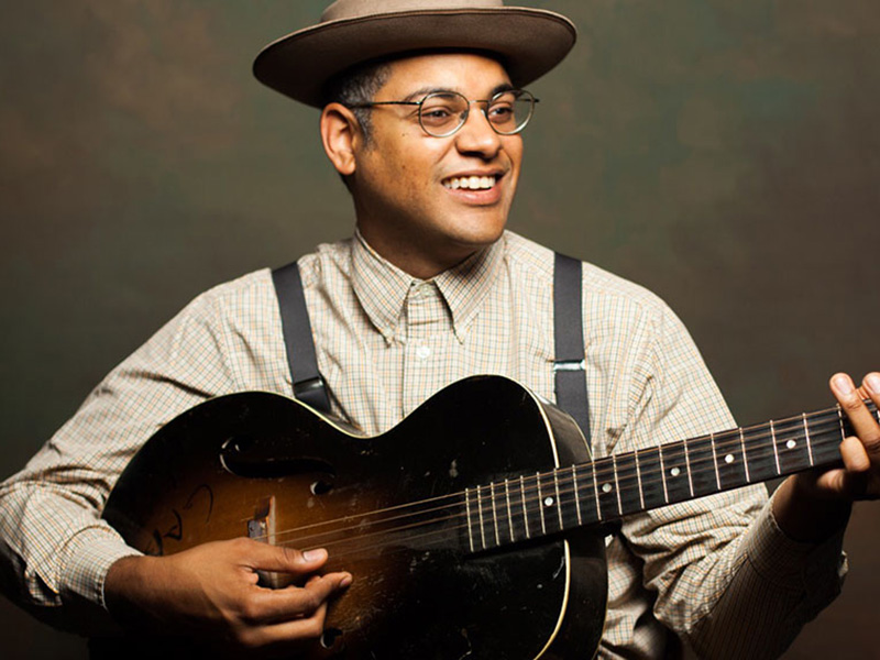 A promotional photo of Dom Flemons holding his guitar