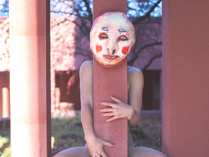 A photo of person's nude body behind a pillar with a mask placed where the head would be