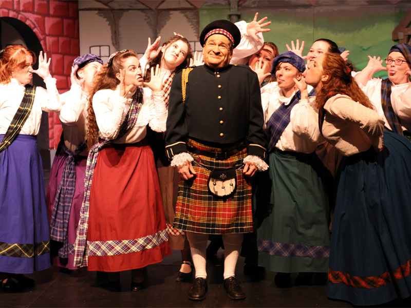 A man stands wearing traditional Scottish kilt, tam and sweater surrounded by women looking adoringly at him. The women are also in traditional Scottish dresses.