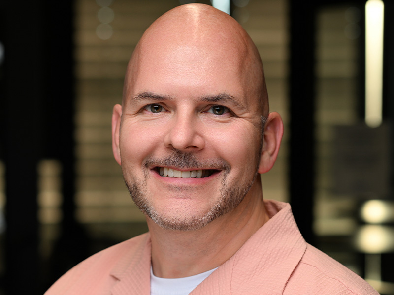 Brian Krywulak in a pink button-down shirt with white tshirt underneath smiling facing forward