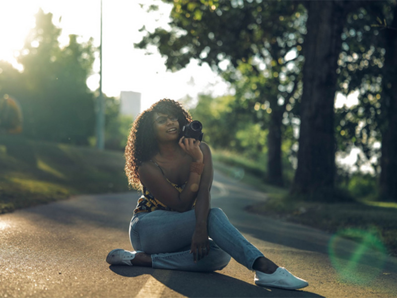 Black woman with curly hair past her shoulders. She is smiling toward the photographer while holding the camera with her leg crossed on the sidewalk lined by trees.