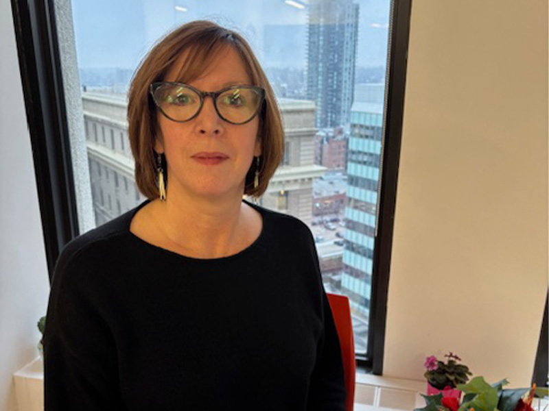 Maureen McNamee sitting in front of a window wearing a black shirt and dark rimmed glasses