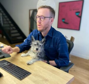A photo of Greg Burbidge holding his dog Scout while sitting at a desk with a keyboard.