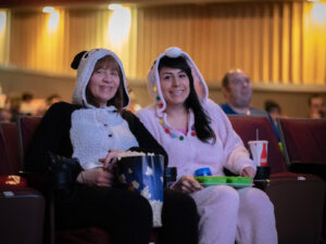 Image of two people sitting in movie theatre wearing onesies and holding snacks