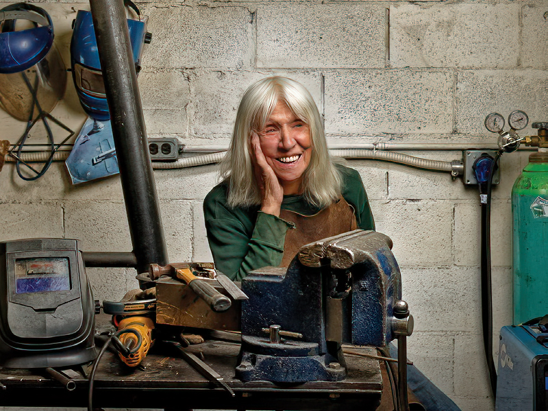 Sculptor Katie Ohe sitting at a table with several tools, and welder's masks hanging on the wall behind her.