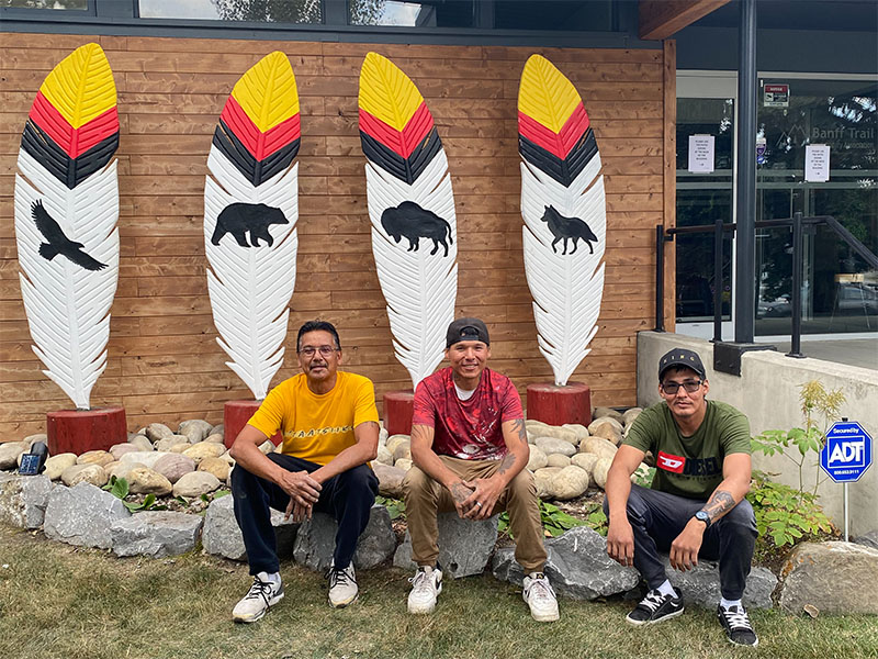 Three men sit in front of four sculptures. Each sculpture is of a feather, and each features a unique animal: an eagle, a bear, a bison and a wolf.