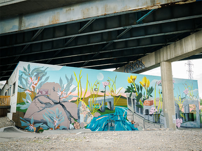A mural featuring flowing rivers and plants growing from concrete is painted on a wall underneath a bridge.