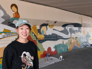 Artist Yiting Hui stands in front her mural, which features a confluence of people and foods from different cultures.
