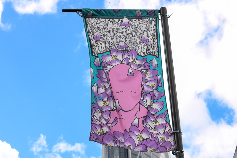 A banner in the wind downtown, featuring a pink person, surrounded by purple flowers.