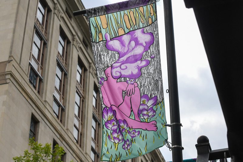 A banner hangs downtown on a pole, featuring a pink person with purple smoke billowing from where their facial features would normally be.