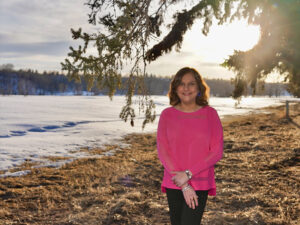 A photo of Kelly Kaur wearing a bright pink top and standing outdoors next to a snow-covered river.