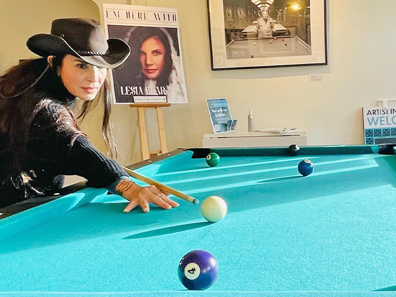 A photo of Lesia Bear wearing a black cowboy hat and standing at a pool table with the cue lined up to strike the white ball.