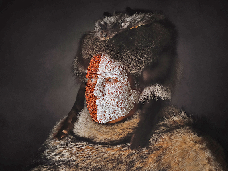A photograph of Half Breed by Marcy Friesen, which shows beaded face that is half white and half brown, wearing a dark fur hat and a lighter brown fur around the neck.