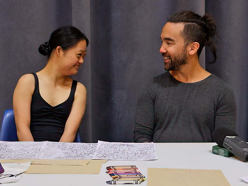 A photograph of dancers Meg Ohsada and Kunji Ikeda sitting side by side and looking at each other behind a table.