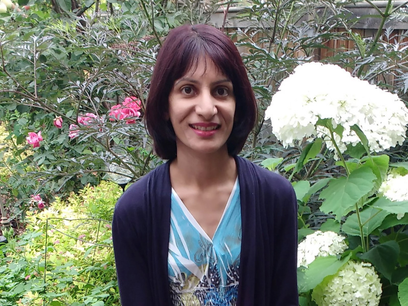 Annie Bhatti wearing a blue shirt and black cardigan standing in a garden in front of green grass and flowers