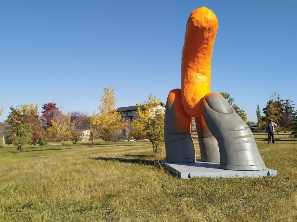 A photo of a large sculpture in a grassy outdoor area. The sculture shows a partial thumb and two fingers emerging from the bottom and holding a giant orange Cheeto.