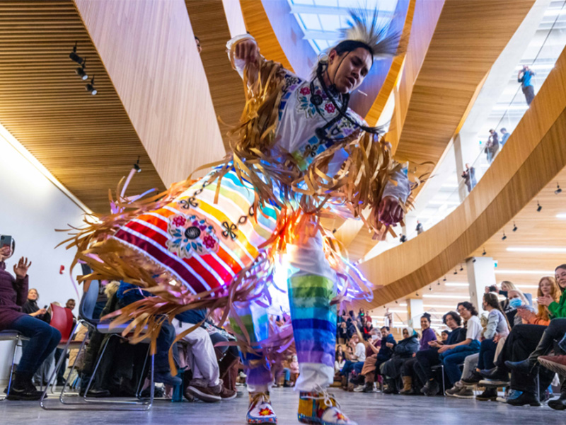 An image of a male Indigenous dancer performing in brightly coloured regalia in the Calgary Public Library