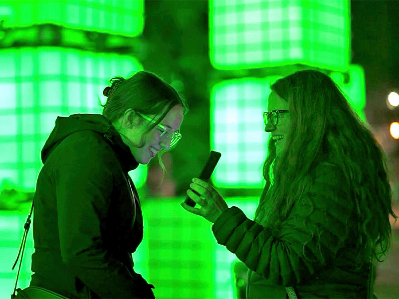 An image of two women facing each other looking at a phone with green lights in the background
