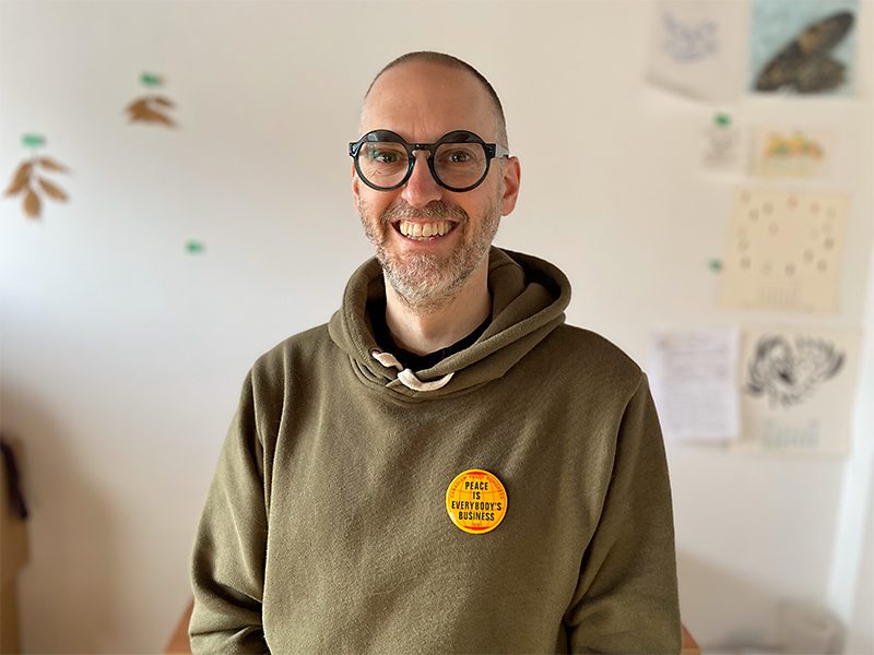 Eric Moschopedis wearing round, black-rimmed glasses and a green hoodie with a yellow button that reads Peace is Everyone's Business.