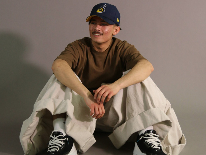 Person sitting on ground wearing brown shirt, ball cap and baggy pants