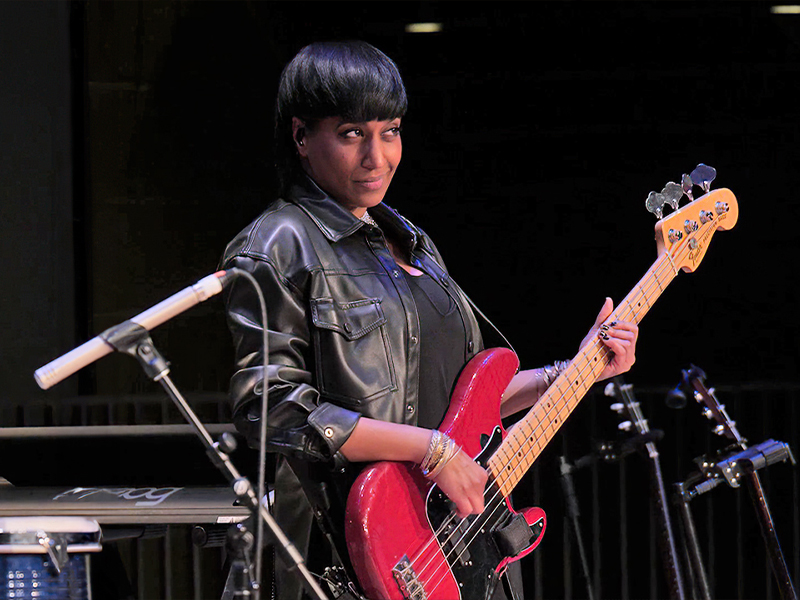 A woman wearing all black holds a red guitar onstage