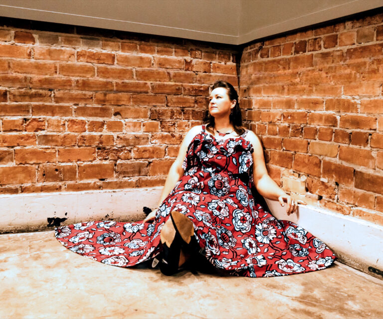 Shone Thistle wearing a floral pattern dress and seated against a brick wall backdrop.