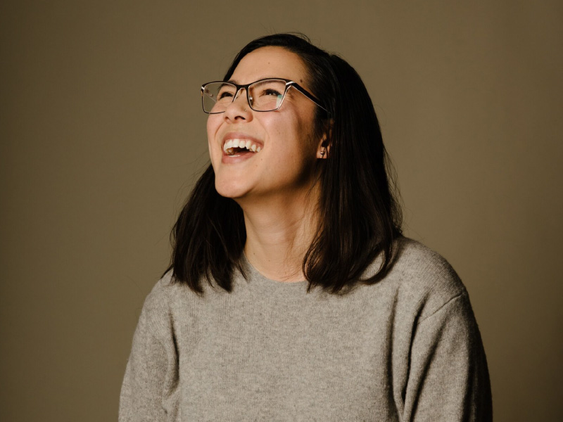 A photo of Jenna looking up and to the left against a brown background. She is laughing with her mouth open. She has shoulder length dark hair and is wearing golden glasses and a fuzzy grey sweater.