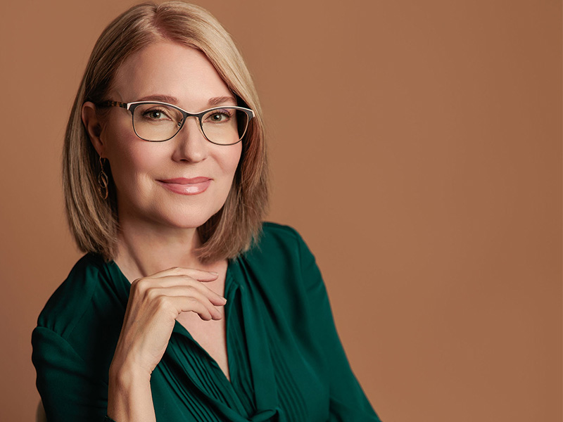 Kirsten Schrader, with blonde hair and glasses and wearing a dark shirt, smiling at the camera.