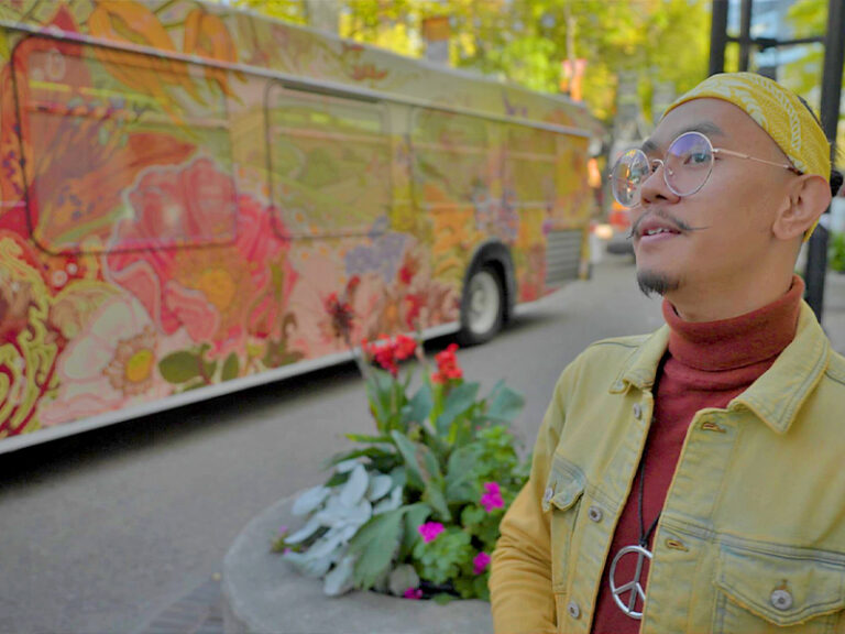 Artist Lyndon Navalta, who has a mustache, goatee and wire-framed glasses, wearing a yellow headband, denim jacket and turtleneck sweater, stands next to his flower-covered art bus at Olympic Plaza.