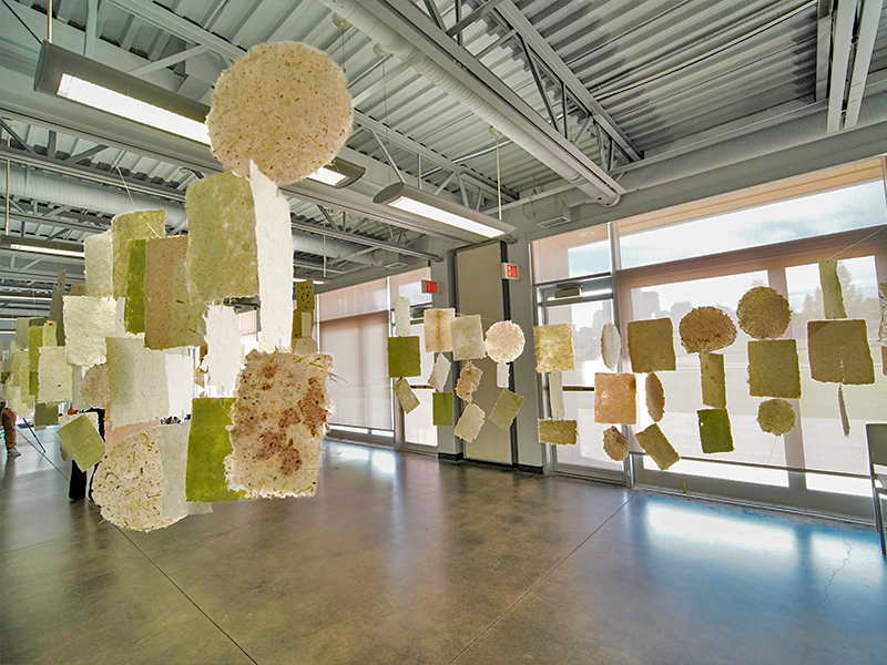 Hanging handmade paper sheets of various sizes and textures are displayed in rows throughout a bright, spacious art gallery with large windows.