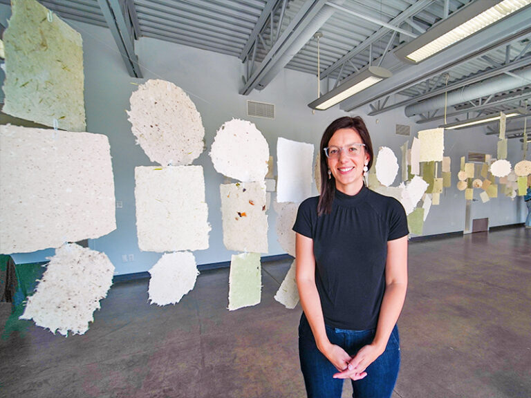 A person wearing glasses and a black top standing smiling in an art gallery, surrounded by large, handmade paper sheets hanging from the ceiling