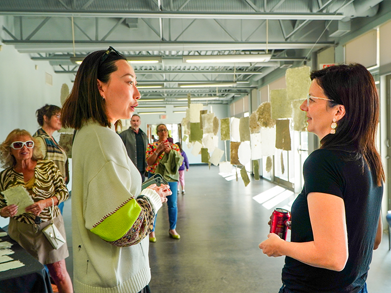 Two women in conversation while others view the hanging handmade paper sheets in the background.