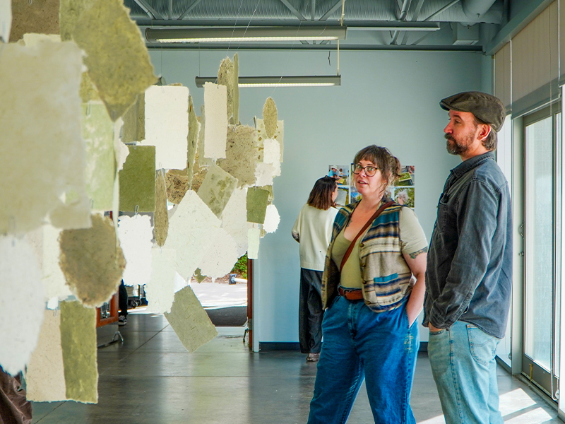 A man and a woman stand in an art gallery, viewing hanging handmade paper sheets, while another person looks at photos on a nearby wall in the background.