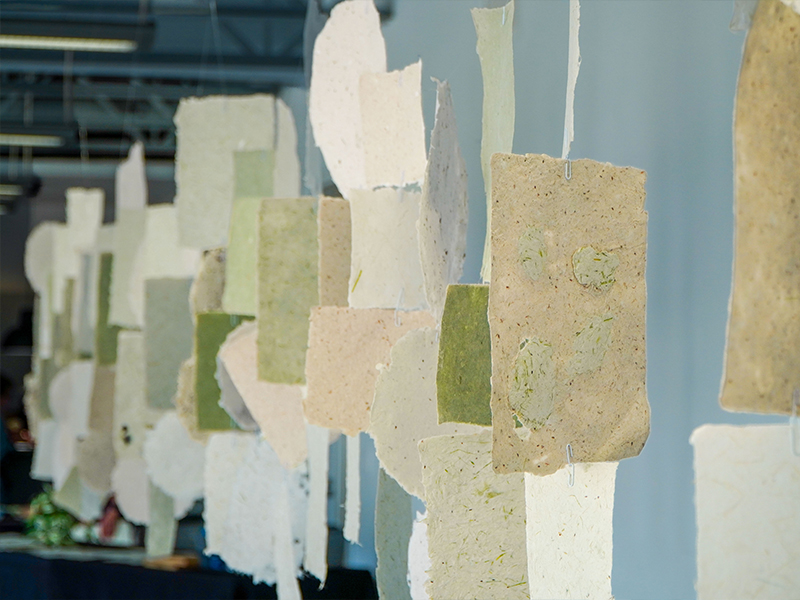 Handmade paper sheets in various shades and textures are hung in rows, creating a layered display in an art gallery.