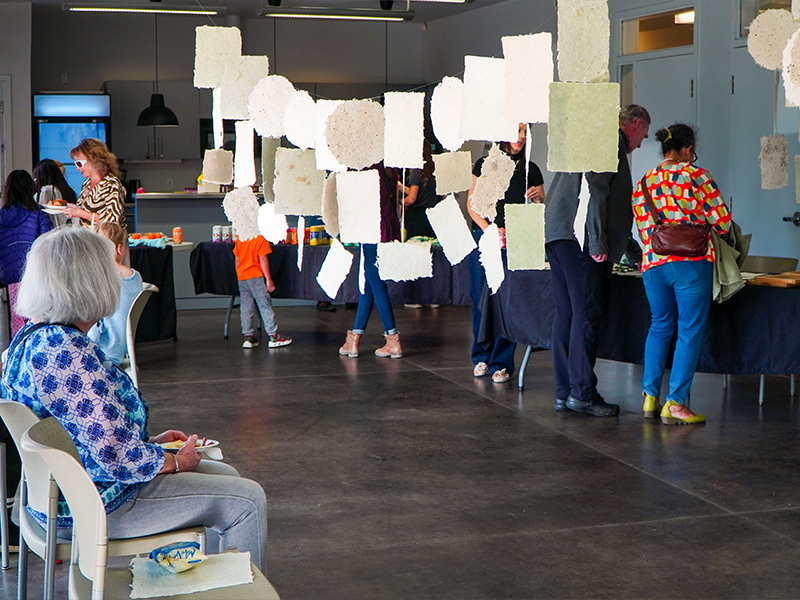 People view hanging handmade paper sheets, with someone seated in the foreground watching.