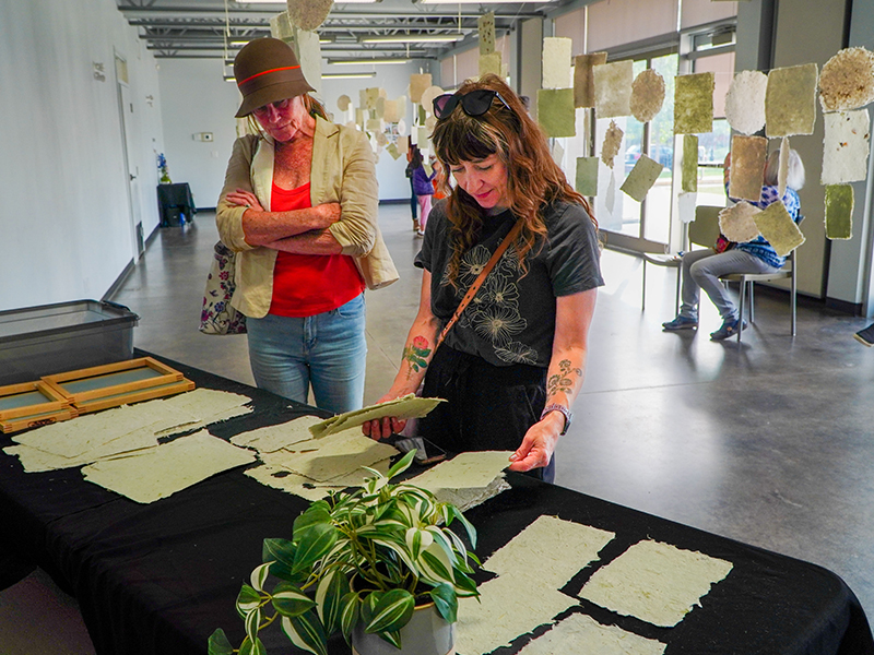 Two women in an art gallery observe sheets of handmade paper displayed on a table, with more paper art hanging in the background.