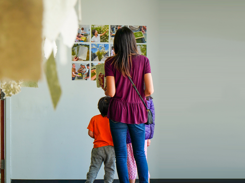 A woman with two children stands in front of a wall displaying photos, viewing the images together.