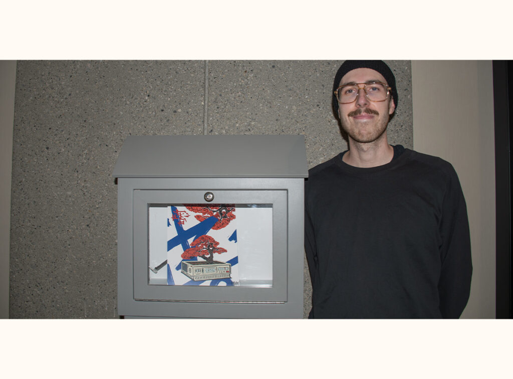 Dylan Nickolet stands beside his mini gallery, displaying an artwork featuring a red tree and architectural elements against abstract blue shapes.