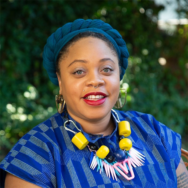 Elisheba Johnson smiling while wearing a blue headpiece, bright patterned blue shirt, large colorful necklace, against a backdrop of greenery.